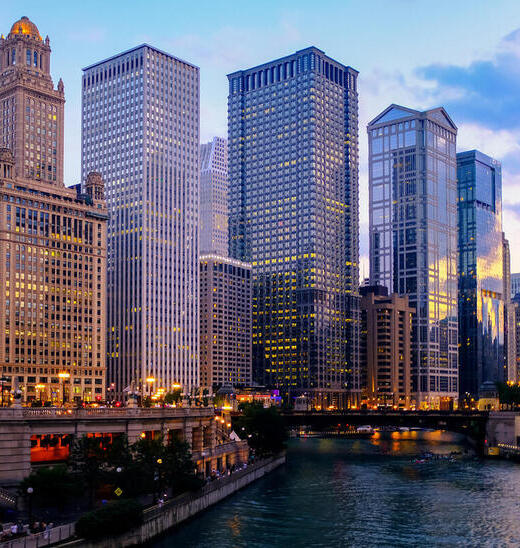 Tall skyscrapers tower over a river at dusk. 