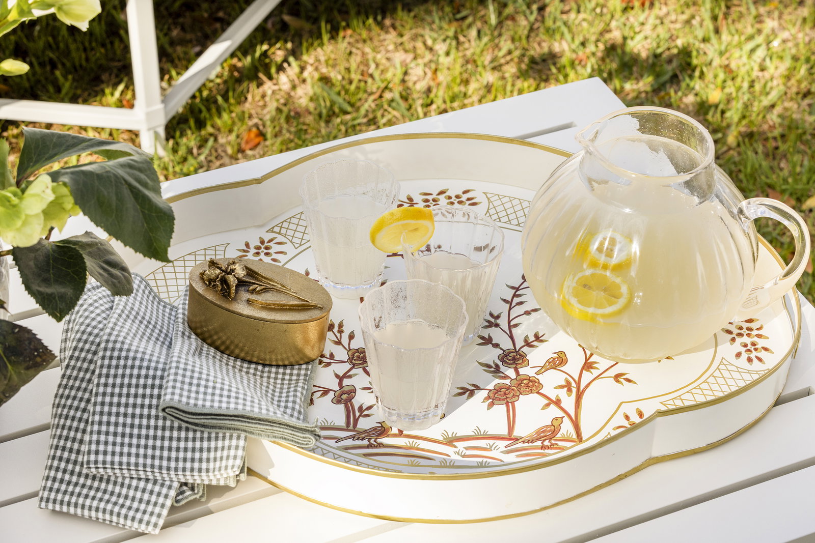 A patterned tray on top of a white coffee table with a pitcher and glasses of lemonade, some green checkered napkins and a gold trinket dish.
