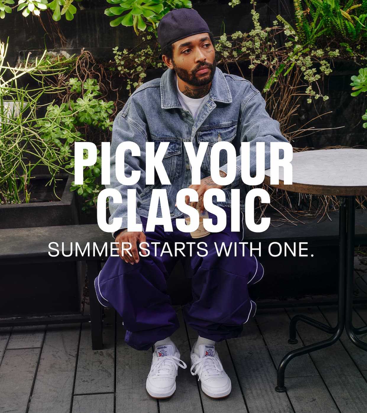Person wearing white Club C 85 sneakers sits outdoors in a denim jacket and purple pants. Text reads: “PICK YOUR CLASSIC. SUMMER STARTS WITH ONE.”