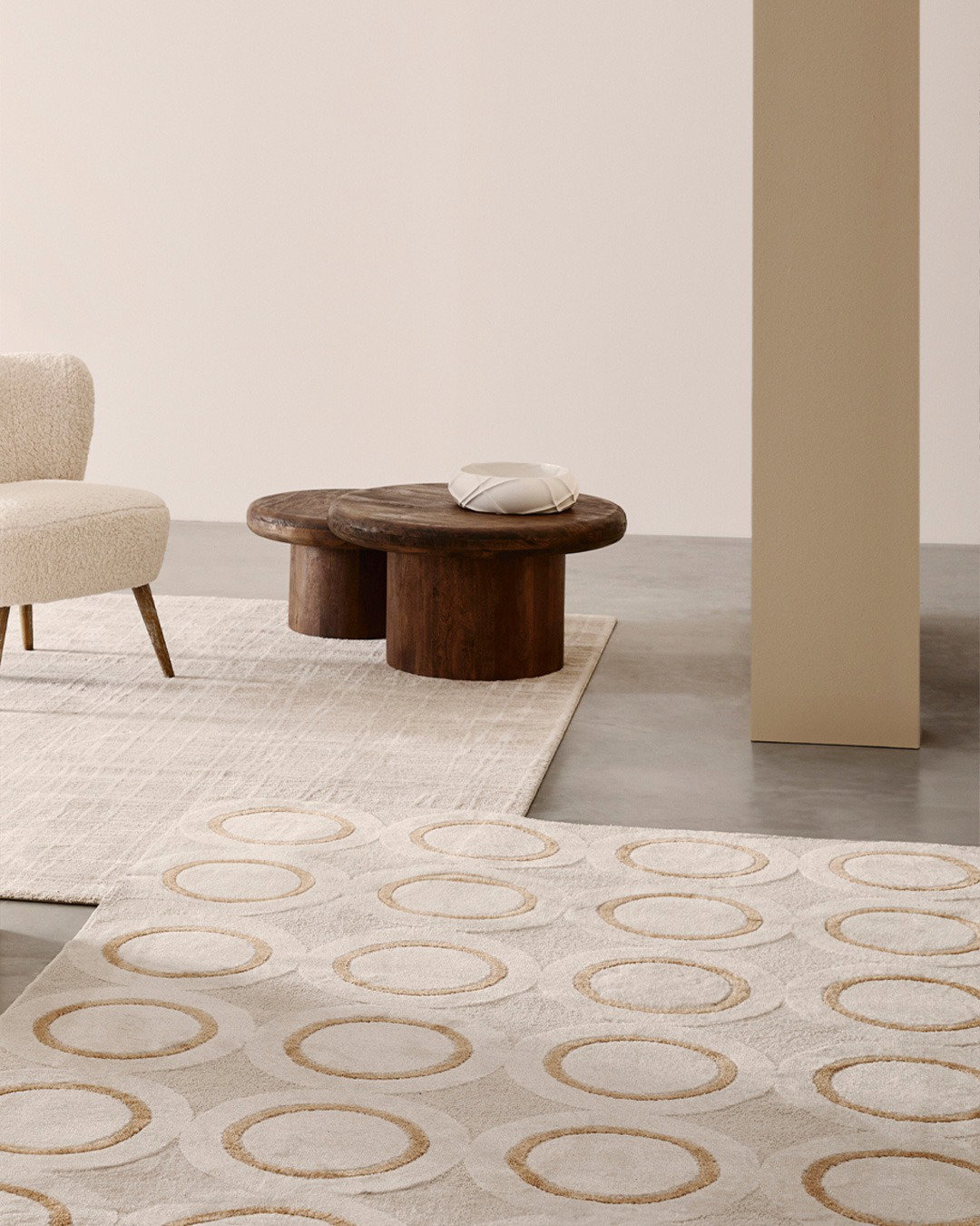 Minimalist living room with cream boucle armchair, two round dark-wood coffee tables on layered neutral patterned rugs over concrete floor