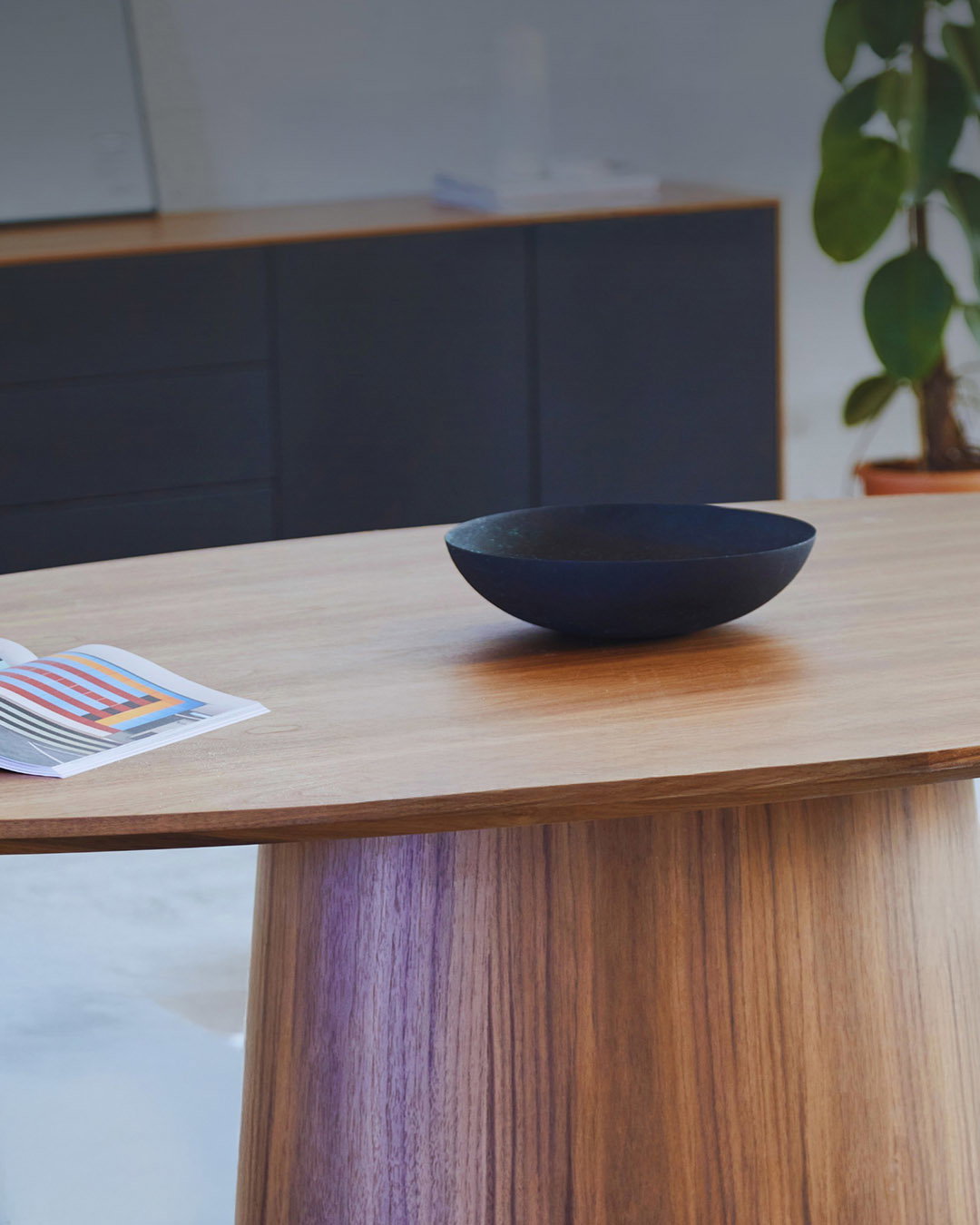 Oak dining table with a simple black decorative bowl and a colourful magazine page, minimalist interior with cabinet and potted plant in background