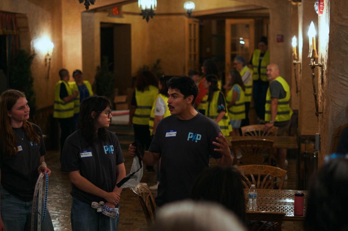 Image of three Powered by People volunteers speaking in a room with tables and chairs. Behind them there is a group of people all wearing high-visibility vests.