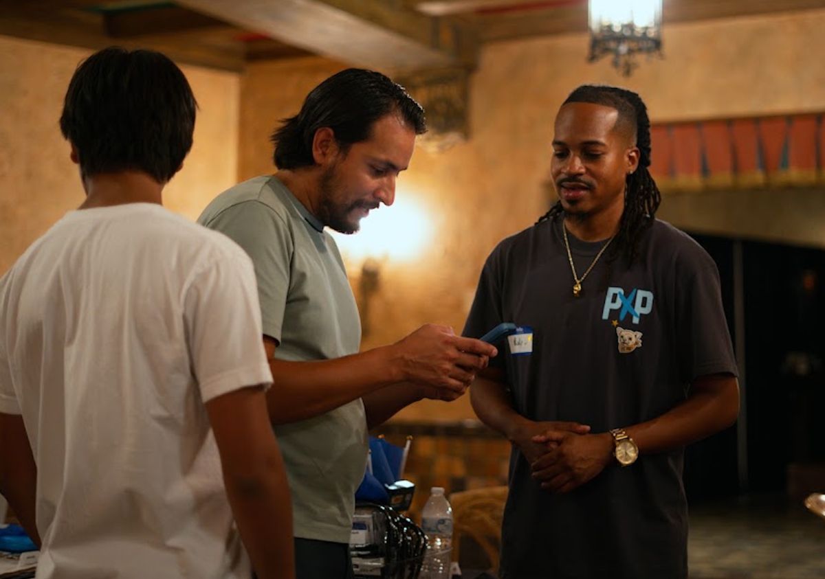 Image of a Powered by People volunteer (right) speaking with a voter wearing a light gray shirt, helping them check their voter registration on their phone. There is another individual facing away from the camera wearing a white tee shirt to the left of the voter.