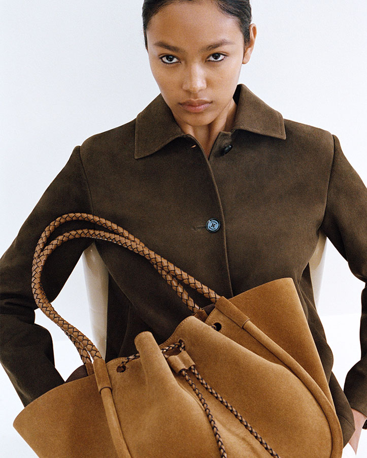 Cropped image of model sitting in chair wearing the Irine Jacket in Suede in Cypress with the Large Ruched Tote in Suede and Braided Calf in Hazlenut in her lap