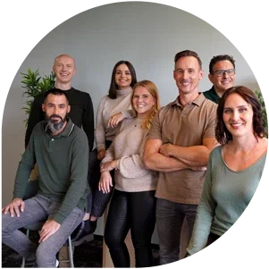 Gruppo di persone in abbigliamento casual, alcune in piedi e altre sedute, con una pianta verde sullo sfondo.