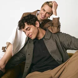 Dos modelos están sentados en una silla marrón. Uno lleva una camisa de cuadros, un polo negro y pantalones beige; el otro lleva un abrigo marrón sobre un vestido blanco.