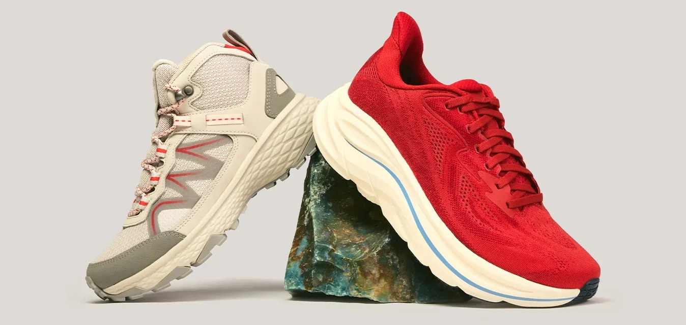 Zapato de senderismo beige y zapato de correr rojo equilibrados sobre una pequeña roca contra un fondo liso.