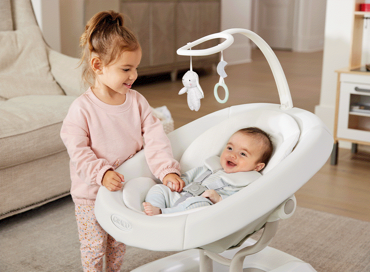 Little girl smiling at a baby in a white baby rocker with hanging toys, both indoors. Baby is buckled and smiling.