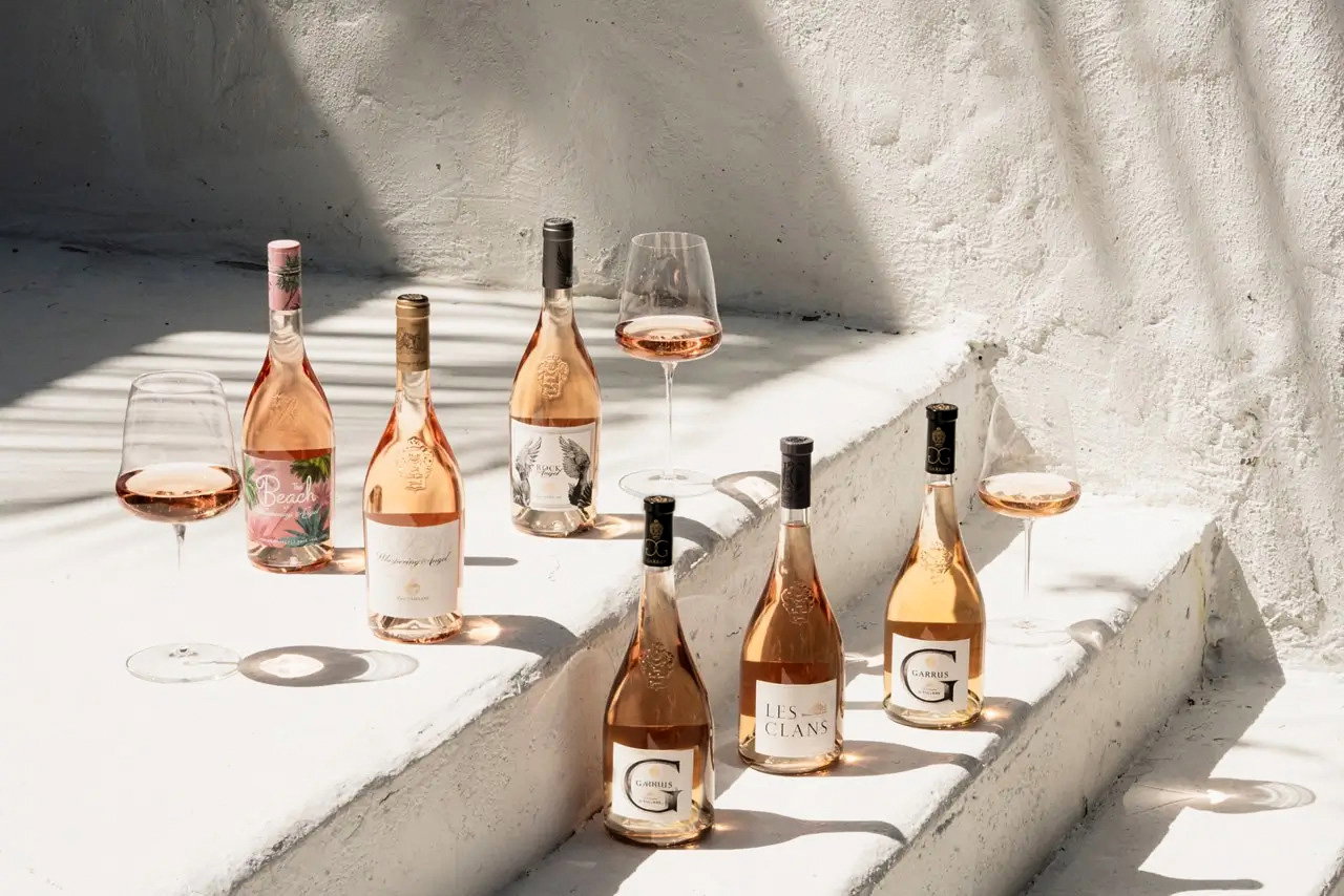 Six rosé wine bottles and three filled wine glasses arranged on sunlit white plaster steps, summer outdoor still life