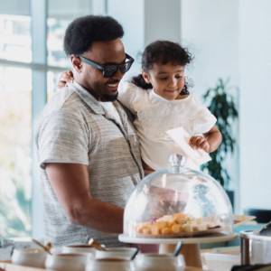 A man holds a young girl while they stand at a counter selecting pastries. The scene appears to be in a bright café or bakery with natural light and modern decor. A glass dome display showcases assorted bite-sized treats in the foreground. Both subjects look happy and engaged, creating a warm, family-oriented atmosphere.