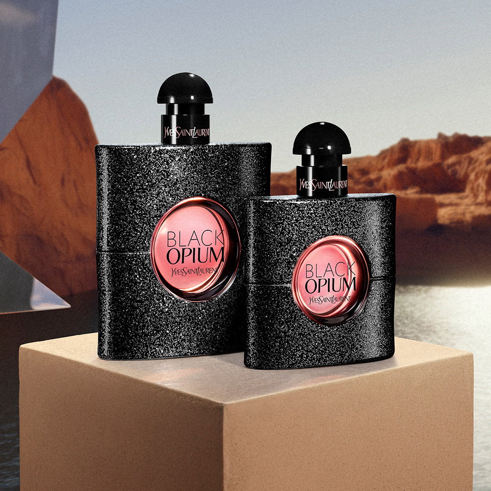 Two glittery black bottles of YSL Black Opium fragrance displayed on a stone block against a desert canyon backdrop.