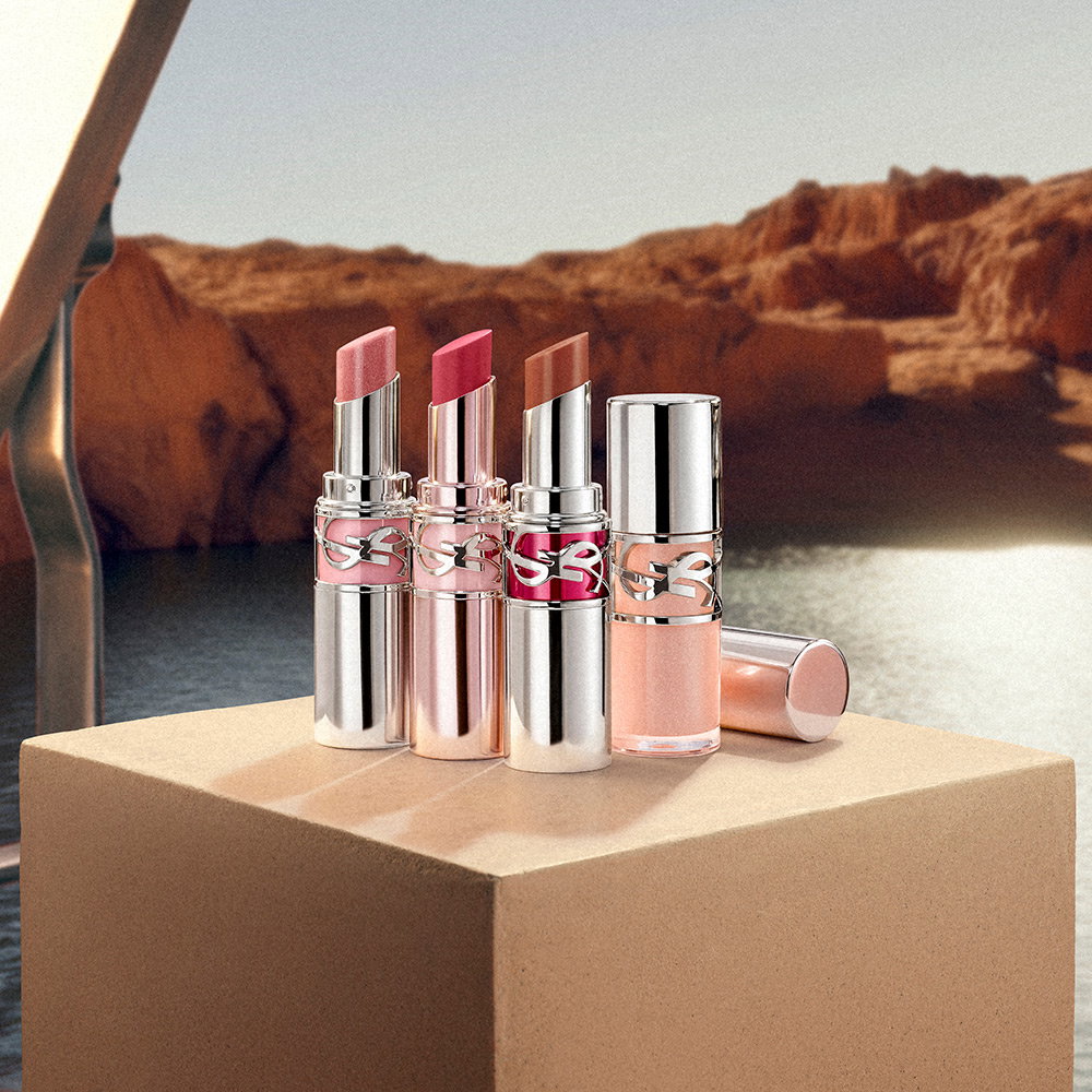 Three YSL lipsticks in shades of pink, berry, and nude alongside a gloss, displayed on a stone block in a desert canyon setting.