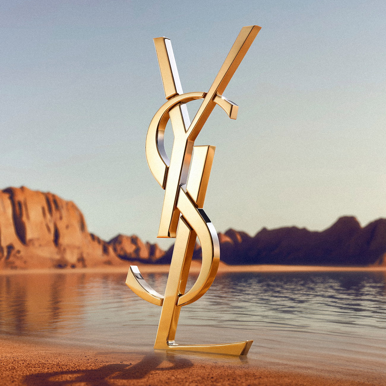 A monumental golden YSL logo standing on a sandy shore with mountains and water in the background.