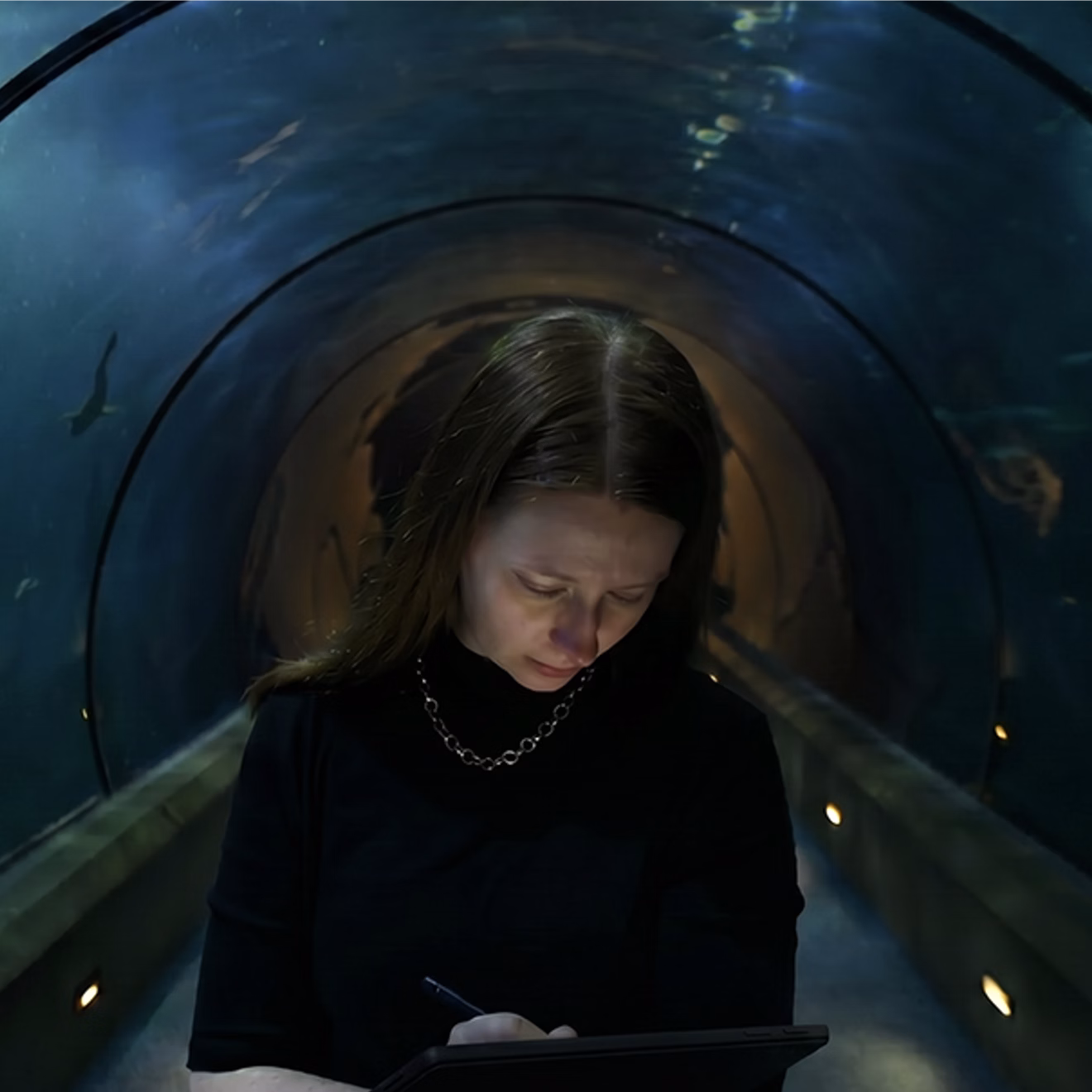 Person standing in an aquarium tunnel, looking down at a tablet and drawing, with sharks and fish swimming overhead.