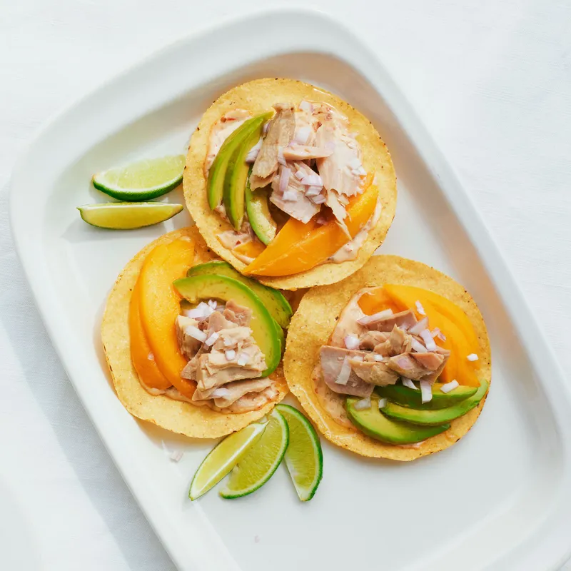 Three tostadas with canned tuna, sliced mangoes, sliced avocados, shallots, and spicy mayo on a platter served with lime wedges and a fourth tostadas on a smaller plate.