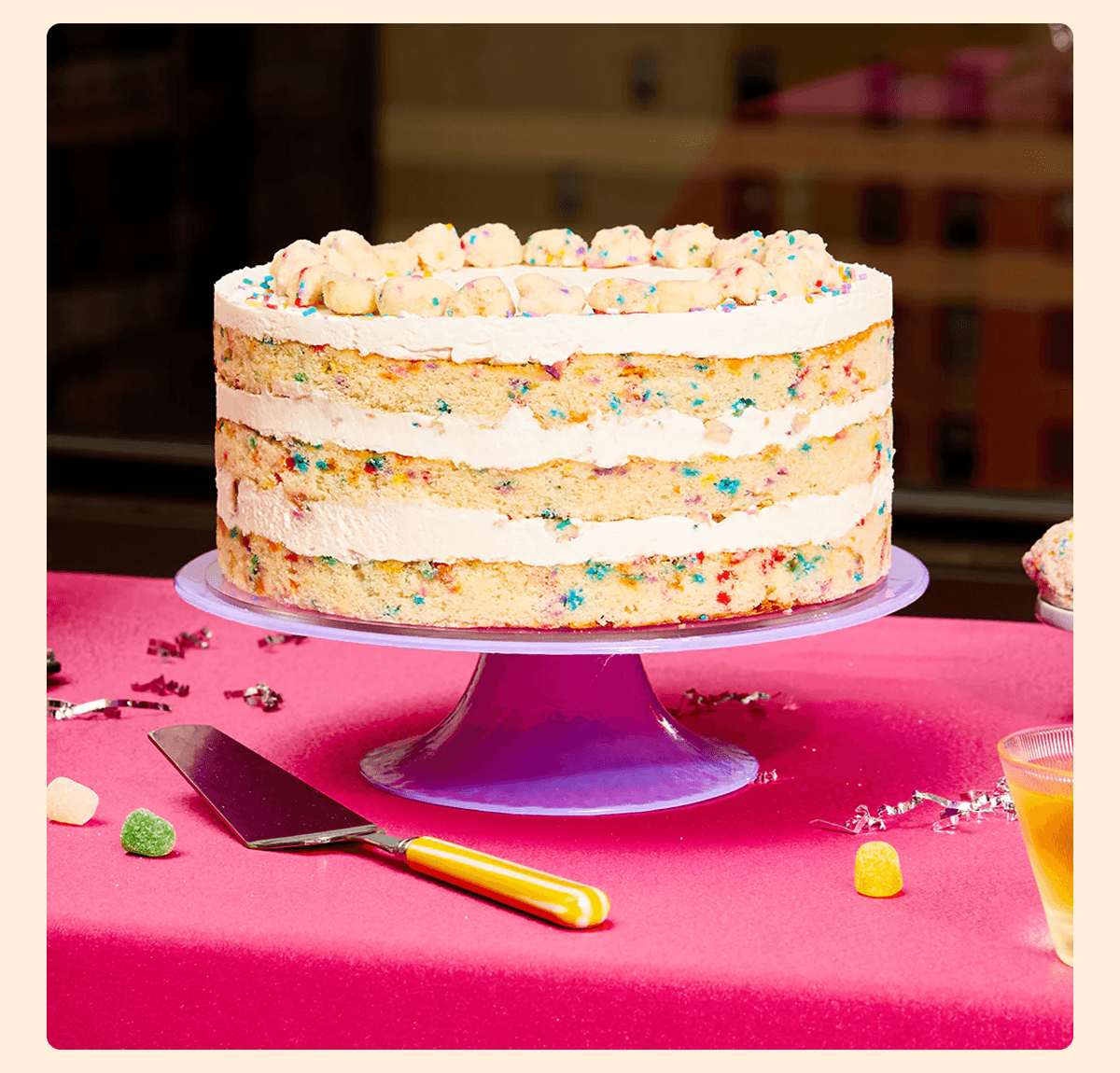 Three-layer confetti birthday cake with white frosting and cake crumbs on a lavender stand, surrounded by a cake server, colorful gumdrops, and silver confetti on a bright pink table.