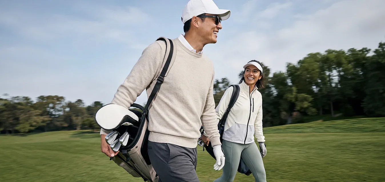 Dos personas caminando por un campo de golf llevando bolsas de golf, vistiendo ropa deportiva informal y sonriendo.