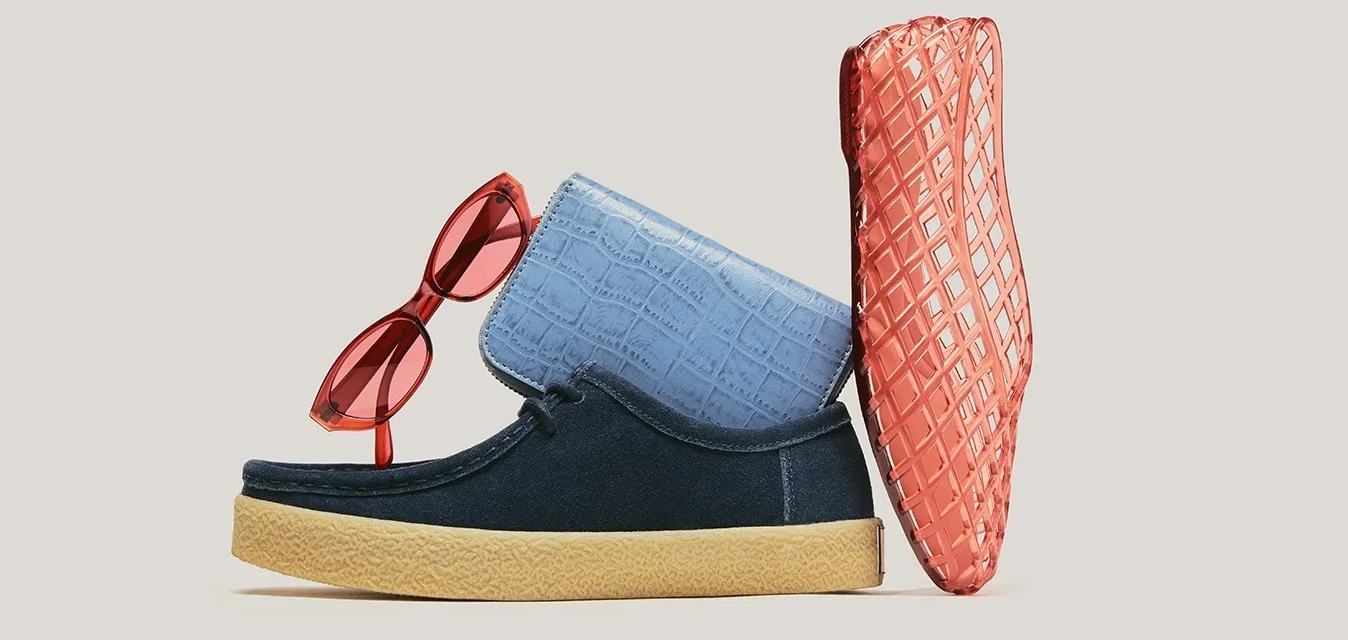 Gafas de sol rojas, zapatilla azul con suela color cuero y bolso coral texturizado dispuestos sobre un fondo beige.