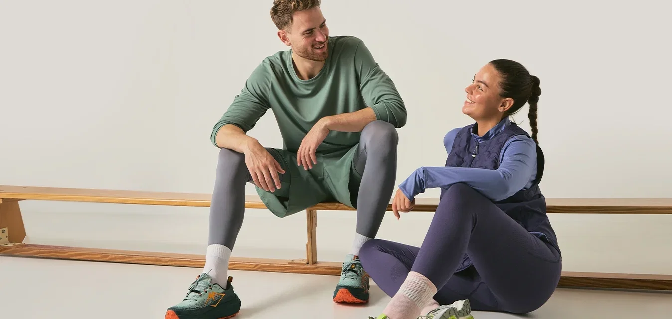 Hombre y mujer en ropa deportiva sentados y sonriendo, descansando sobre un banco de madera y cerca de él en un espacio brillante y minimalista.