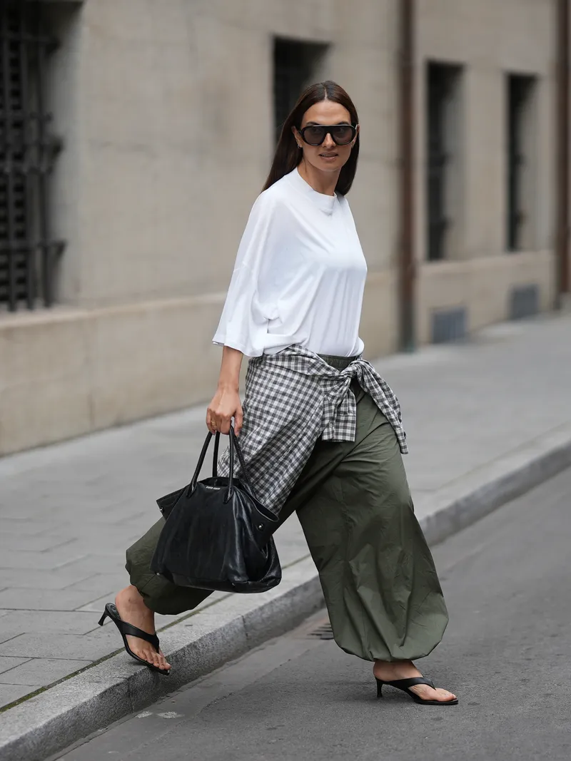 how to style balloon pants - MUNICH, GERMANY - JUNE 16: Anna Winter is seen wearing an oversized white cotton T shirt from Jil Sander; a checked shirt from Pull&Bear tied around the waist as an additional layer; olive green balloon trousers from Herskind; black leather kitten heel thong mules from ROTATE; a vintage black leather handbag from Miu Miu; a gold Cartier Love Ring; aviator sunglasses with dark tinted lenses from Prada on June 16, 2026 in Munich, Germany. (Photo by Moritz Scholz/Getty Images)