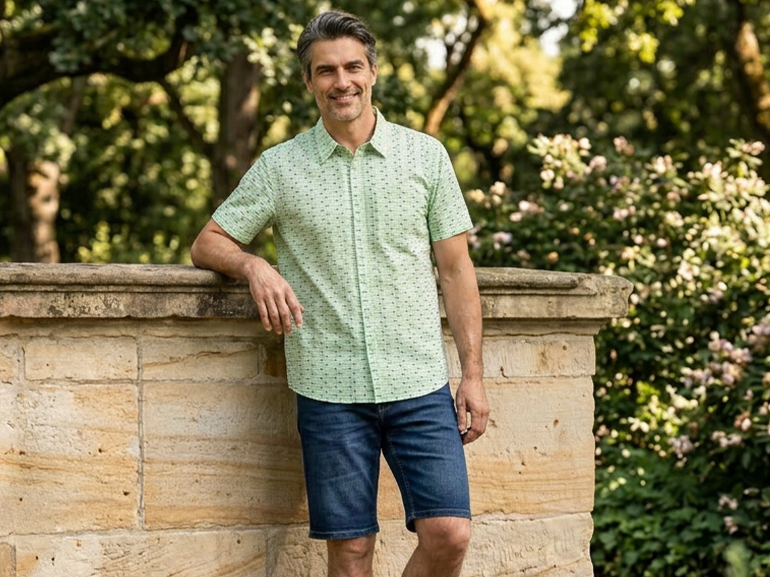 Ein Mann mit grauem Haar lehnt an einer Steinmauer im Freien, trägt ein hellgrünes, gemustertes Hemd und blaue Jeansshorts.