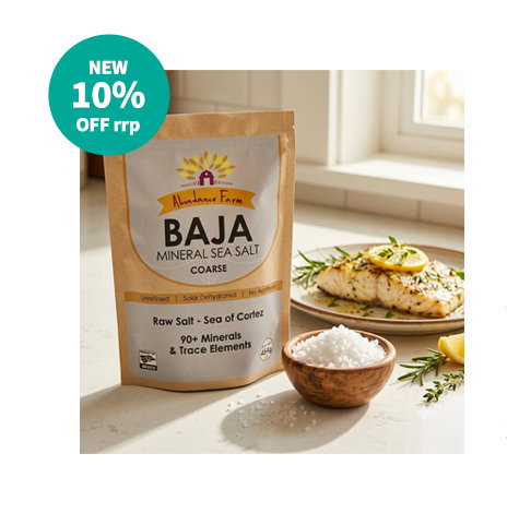 A pouch of BAJA Gold Mineral Sea Salt stands on a kitchen bench next to a small wooden bowl of coarse sea salt, and a white fish dish topped with sliced lemons and herbs.