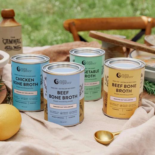 A range of Nutra Naturals organic bone broths in tins, including Beef, Chicken and Vegetable varieties, displayed outdoors on rustic picnic table like setting. Deal includes Broth ranges, collagen and more.