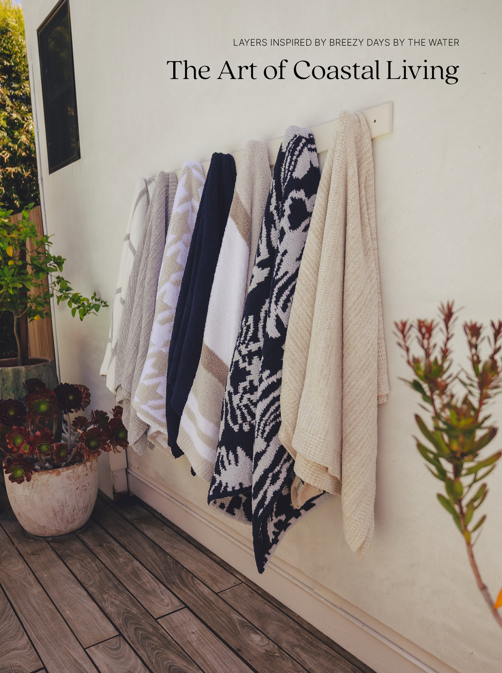 A collection of plush Kashwére throws in coastal-inspired patterns and neutral shades hangs on an outdoor wall rack beside potted plants, creating a relaxed patio setting. The headline reads, "Layers Inspired by Breezy Days by the Water" above "The Art of Coastal Living."
