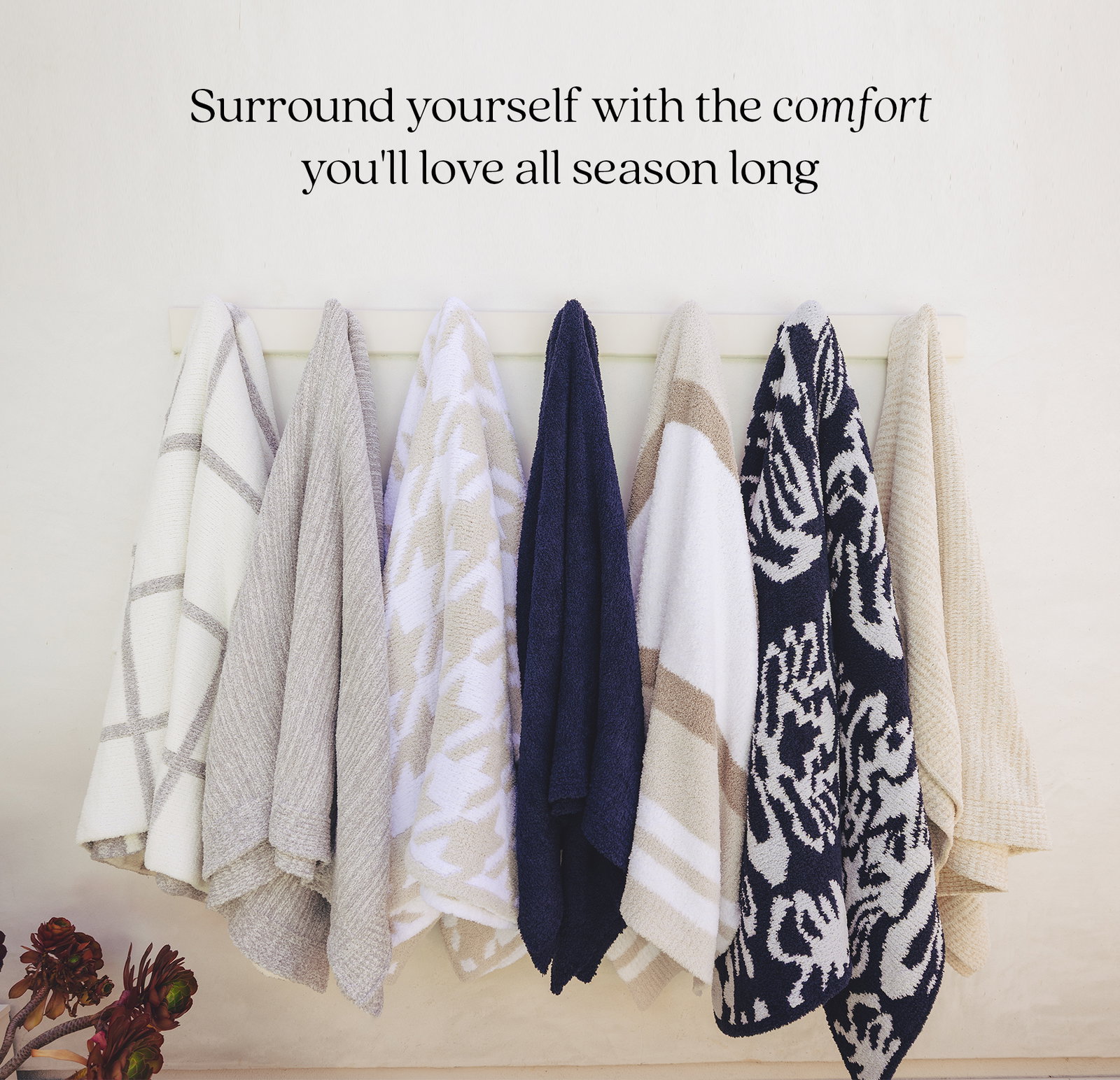 Seven plush Kashwére throws in a mix of coastal-inspired patterns and solid neutrals hang on a wall rack, featuring windowpane, heathered, geometric, striped, damask, and textured designs in cream, beige, and navy. The headline above reads, "Surround yourself with the comfort you'll love all season long."
