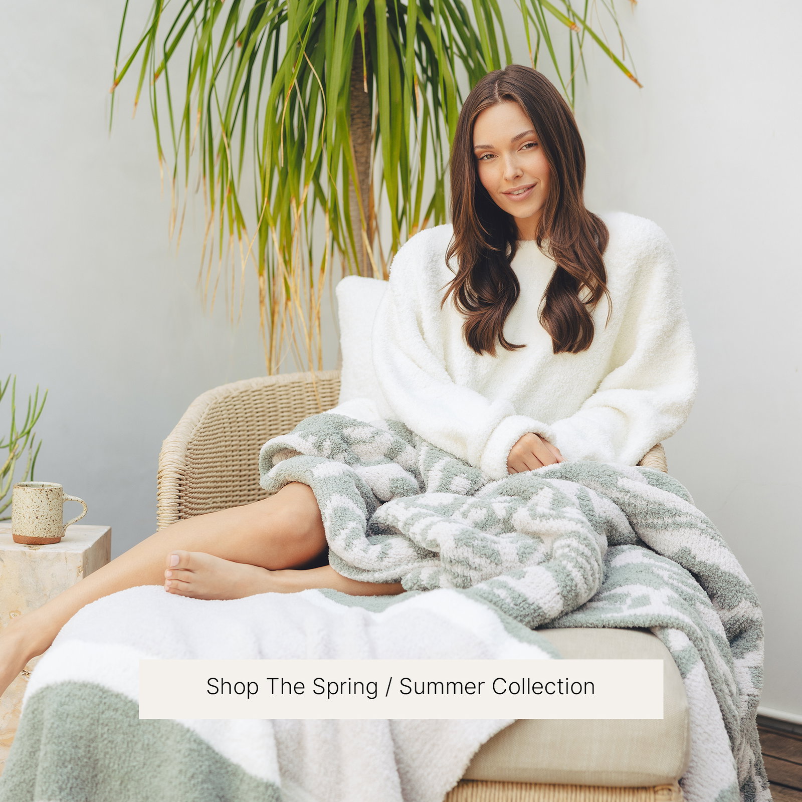 Woman relaxing in a wicker lounge chair wearing a cozy white sweater, wrapped in a sage green and white patterned Kashwére throw. A potted palm and ceramic mug create a bright, airy setting. A "Shop The Spring / Summer Collection" button appears below the image.