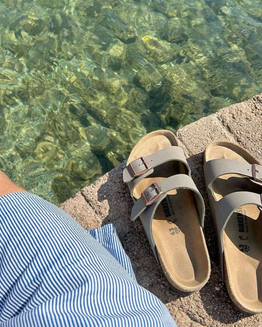 Tan cork sandals and striped blue shorts on a stone pier overlooking clear green seawater
