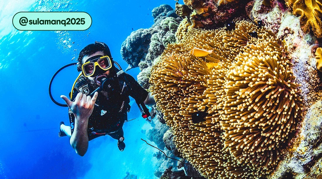 Image description: A scuba diver throwing a shaka underwater next to coral, photo taken by @sulamanq2025