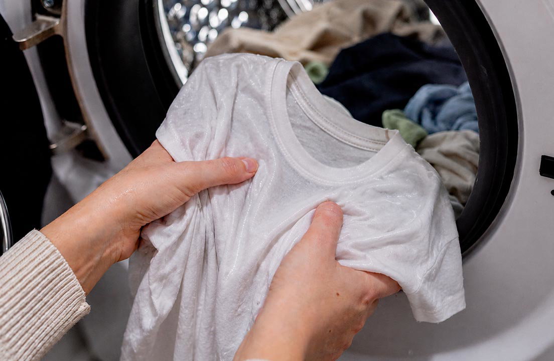 Hands hold a damp white shirt in front of an open dryer filled with clothes