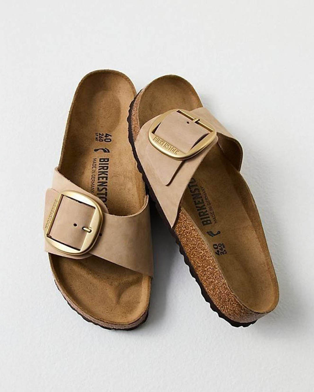 Beige single-strap Birkenstock sandals with large gold buckle and contoured cork footbed, top-down view on pale background
