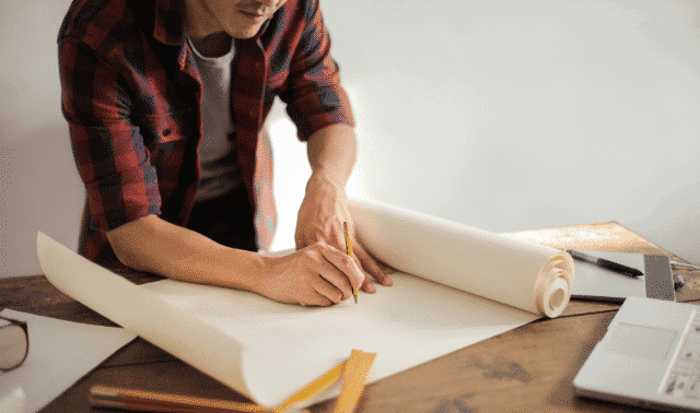 A person in a plaid shirt sketches on a large roll of paper on a wooden table, with drafting tools and a laptop nearby.
