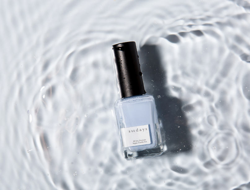 Bottle of pearlescent blue nail polish standing in rippling water with shimmering reflections.