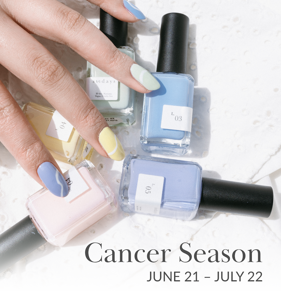 Hand with a pastel blue manicure holding four nail polish bottles in blue, mint, and pearlescent shades against a white wavy background.