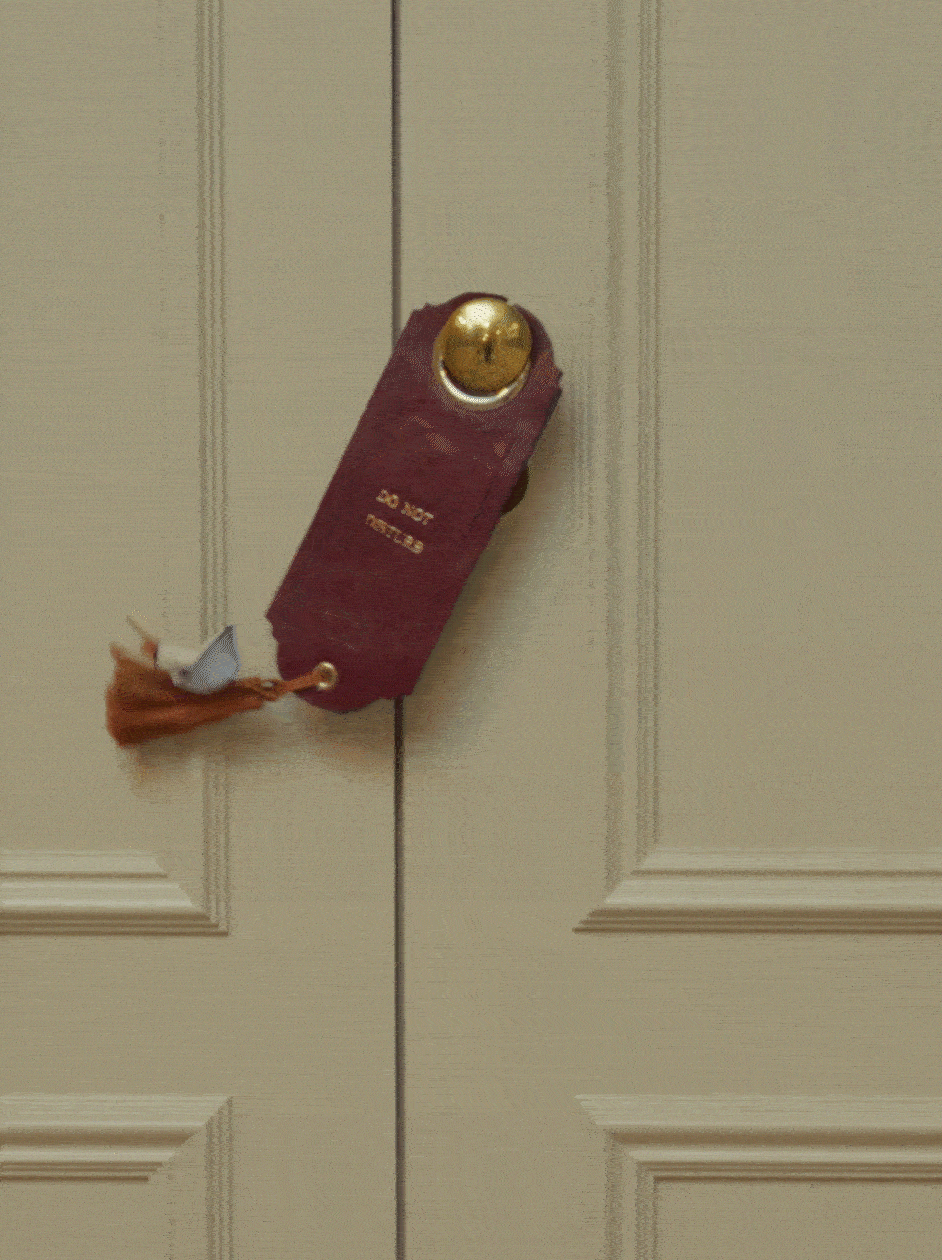 Do-not-disturb-Türschild aus dunkelrotem Leder mit goldener Prägung „Do Not Disturb“, hängt am goldenen Türknauf einer cremefarbenen Kassettentür, mit Quaste in Rostbraun und Weiß verziert.