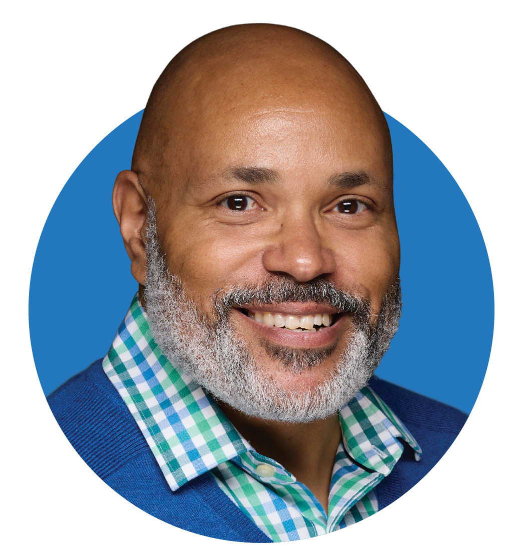 A smiling man with a beard and mustache wearing a blue sweater and a green and white checkered shirt.