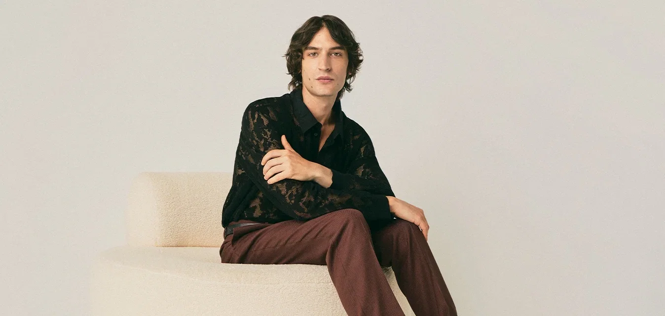 Jonge man zittend op een ronde beige poef, gekleed in een zwart kanten shirt, bordeauxrode broek, gemusterde sokken en bruine leren schoenen in een minimalistische kamer.