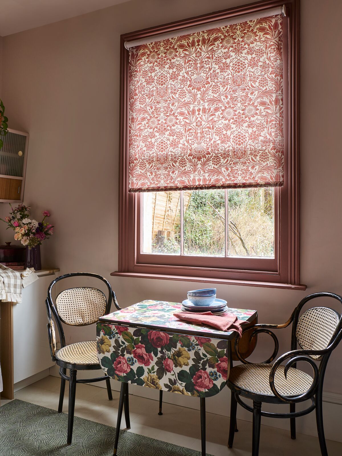 Window with patterned Roman blind filtering light above a small dining table in a cosy kitchen nook with soft pink walls.