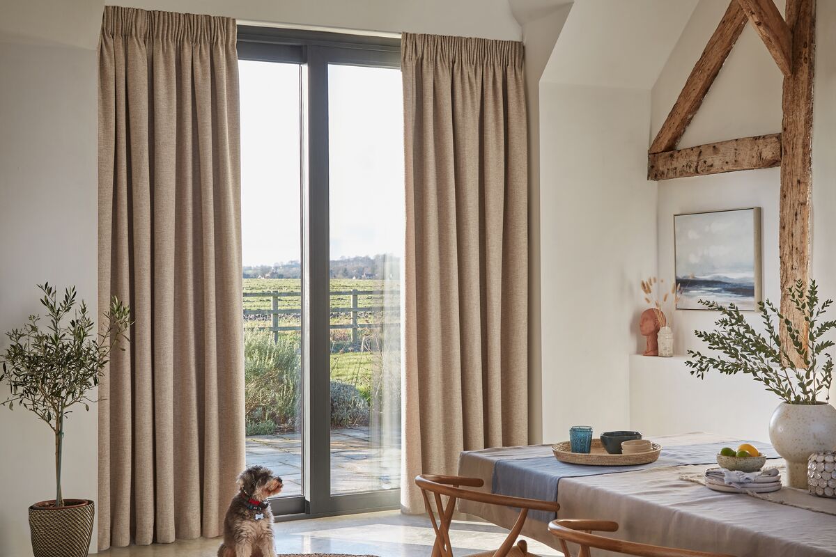 Beige curtains frame large sliding glass doors to countryside, daylight filling dining area with table, chairs and a dog on a woven rug.