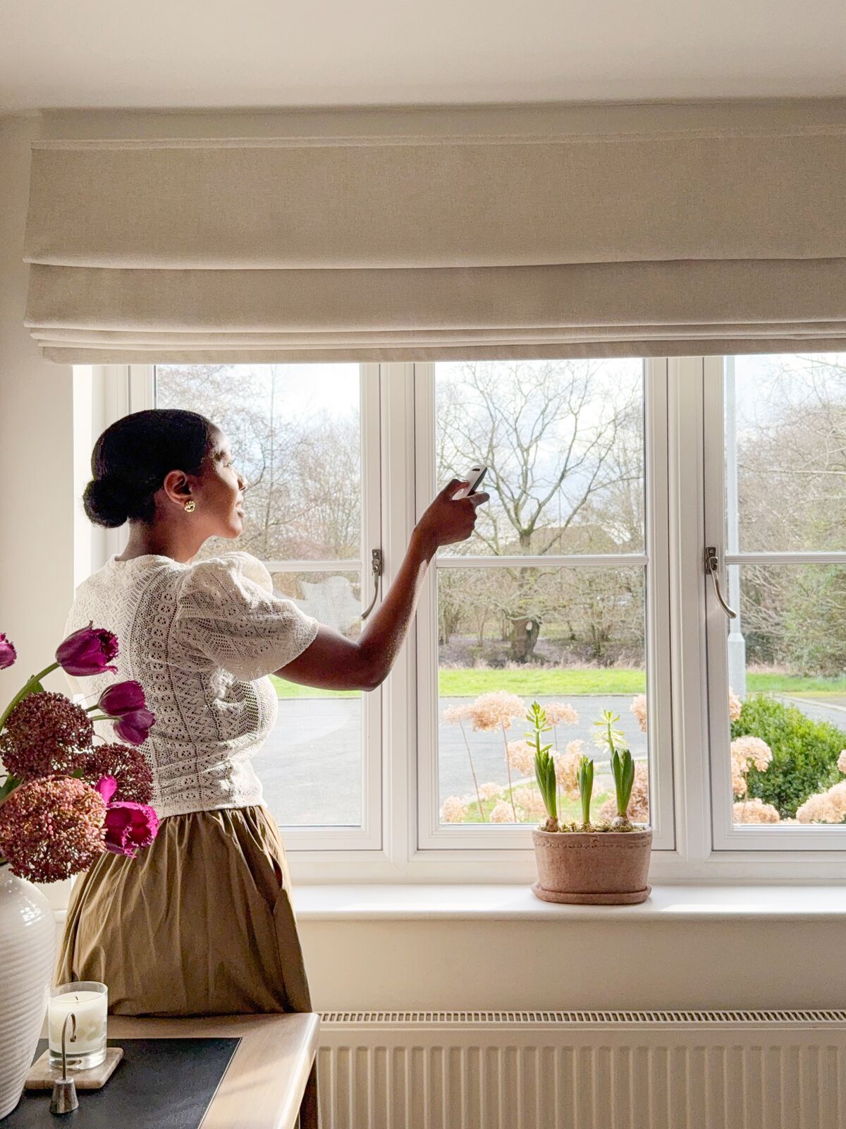 Person adjusting a Roman blind over a bright window with garden view, flowers on the sill and soft neutral décor.