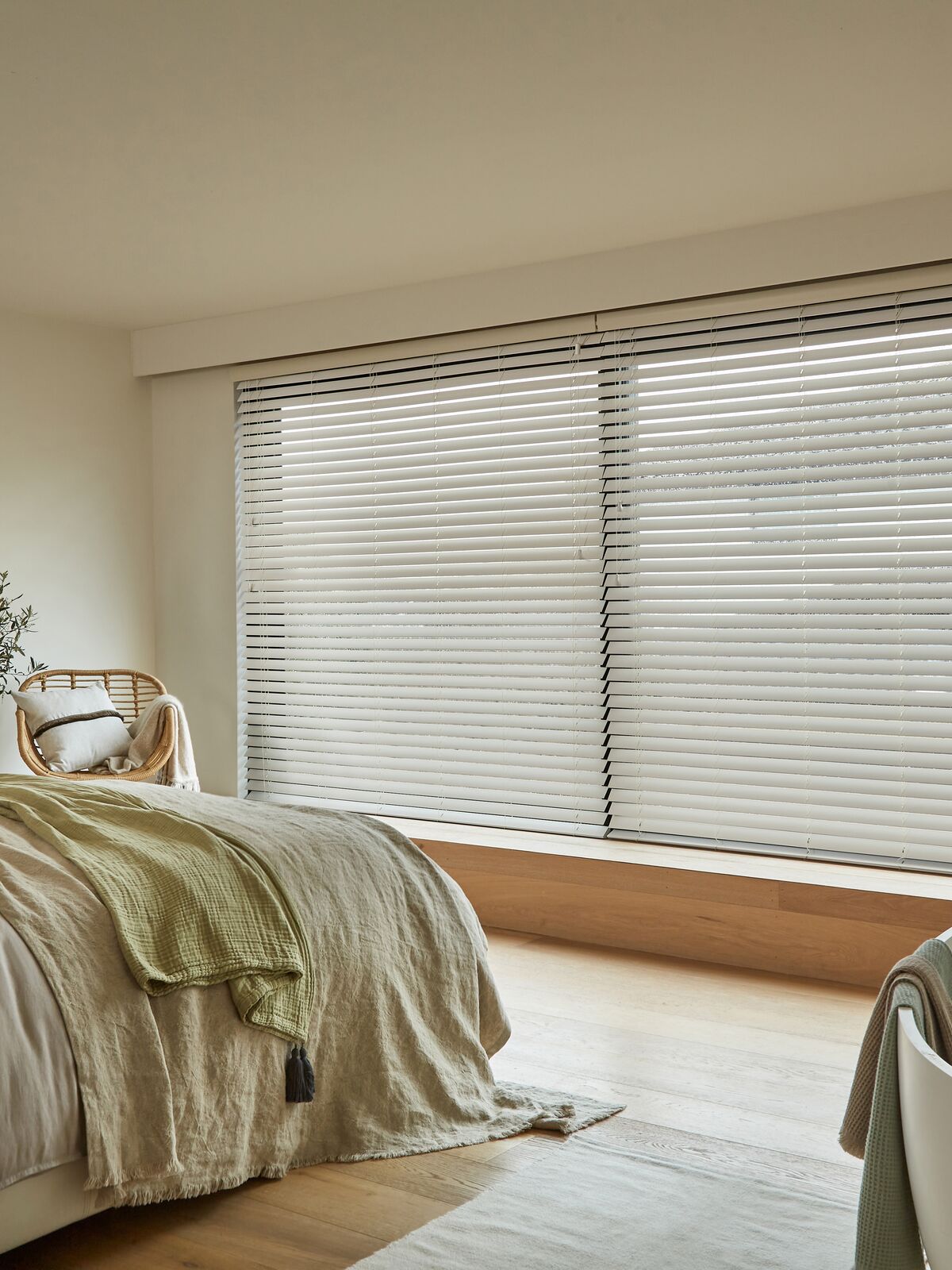 Bedroom with wide window fitted with horizontal blind, soft daylight filtering across neutral bedding and minimalist décor with wooden accents.