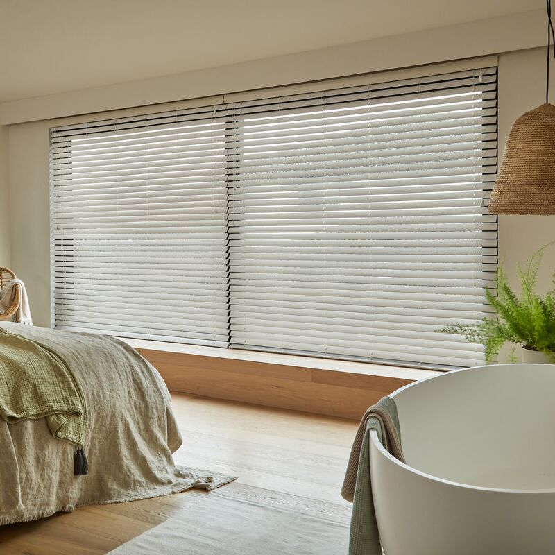 Bedroom with wide window fitted with horizontal blind, soft daylight filtering across neutral bedding and minimalist décor with wooden accents.