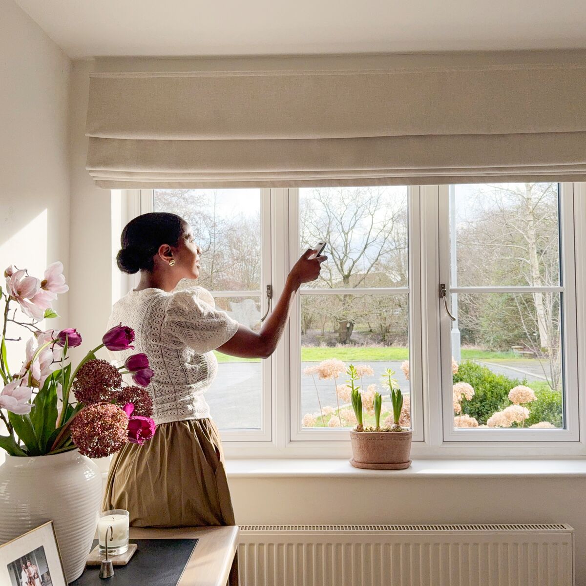Person adjusting a Roman blind over a bright window with garden view, flowers on the sill and soft neutral décor.