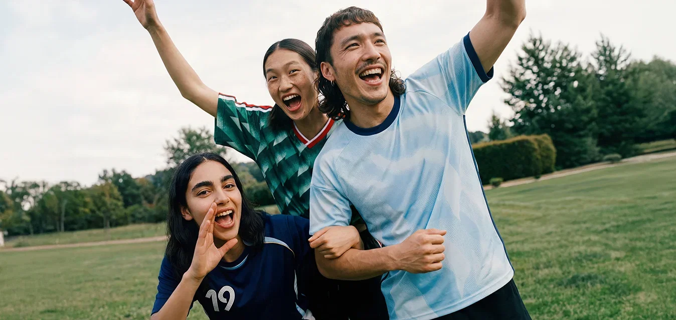 Tre giovani adulti in abbigliamento sportivo applaudono e sorridono all'aperto in un parco erboso con alberi sullo sfondo.