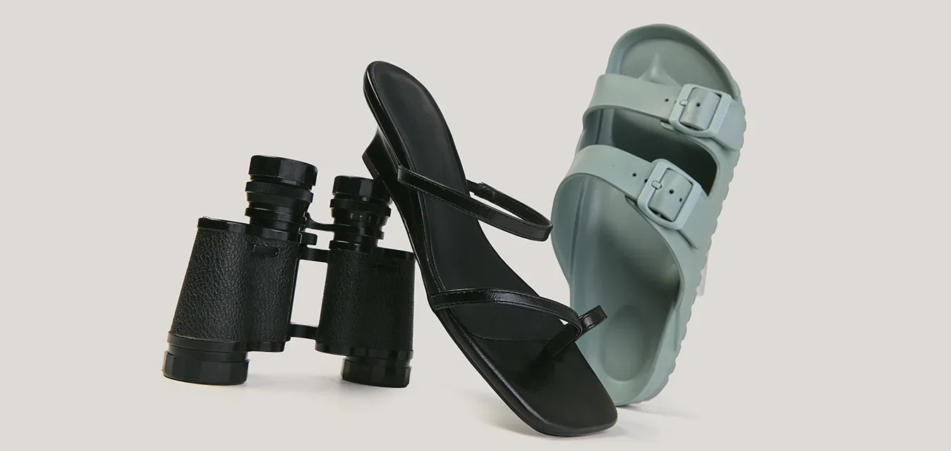 Binocolo nero, sandalo nero con tacco alto e sandalo scivolo verde chiaro con doppio cinturino disposti uno accanto all'altro su uno sfondo semplice.
