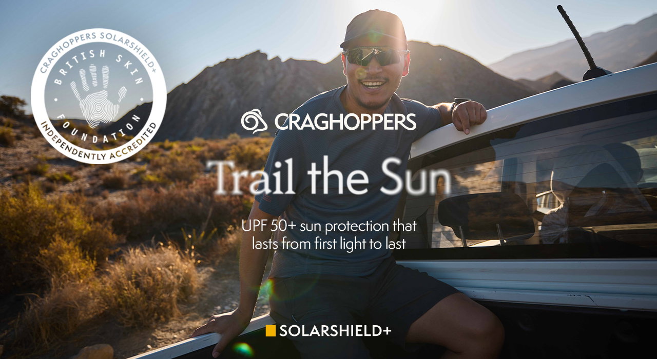 Person sitting on a vehicle in a sunny, mountainous outdoor setting with warm light and lens flare. Branded Craghoppers banner reads “Trail the Sun” with “UPF 50+ sun protection that lasts from first light to last,” plus SolarShield+ and British Skin Foundation accreditation badge.