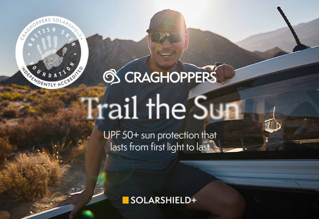 Person sitting on a vehicle in a sunny, mountainous outdoor setting with warm light and lens flare. Branded Craghoppers banner reads “Trail the Sun” with “UPF 50+ sun protection that lasts from first light to last,” plus SolarShield+ and British Skin Foundation accreditation badge.